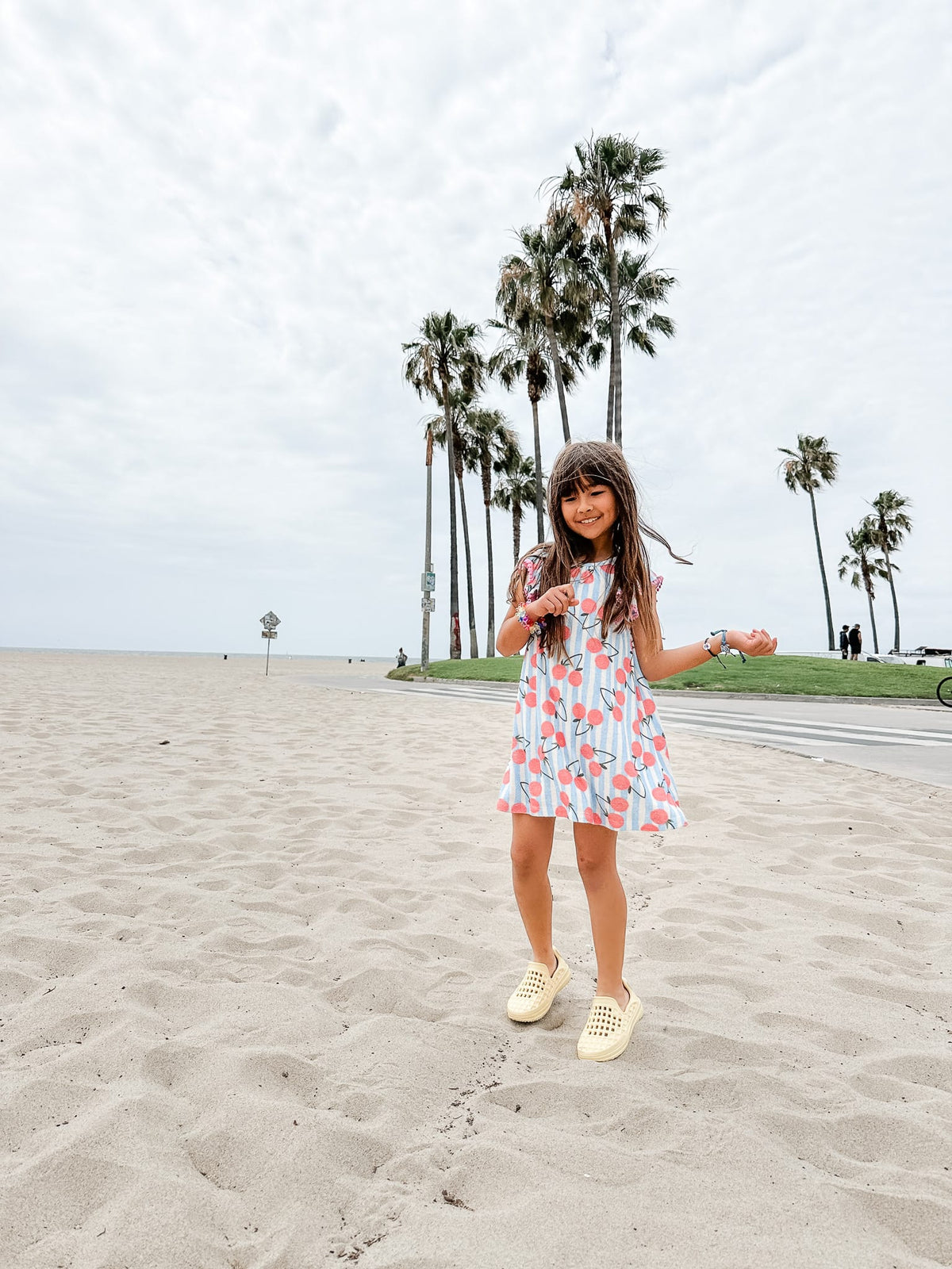 Young girl in a floral dress standing on a sandy beach with palm trees in the background.